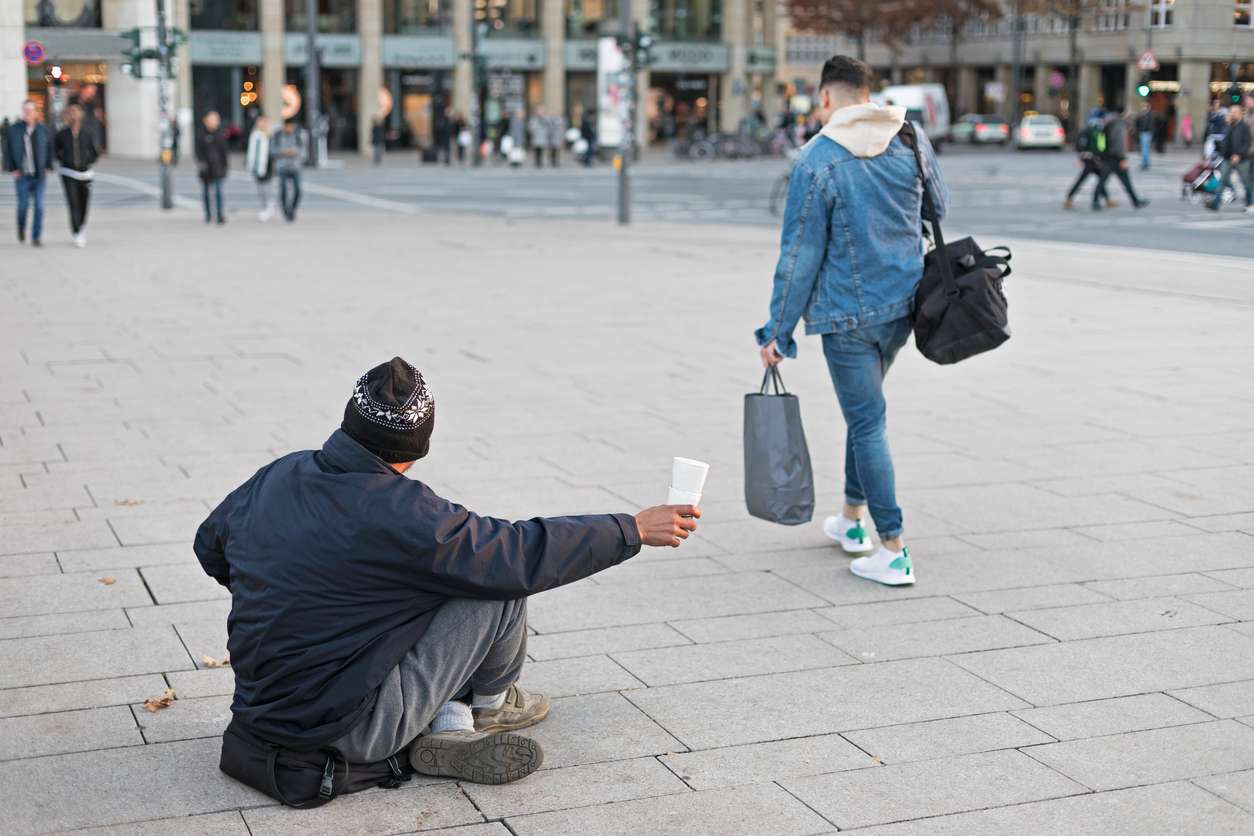 un homme assis sur une place dans une ville mendie en tendant un gobelet